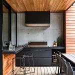 a-contemporary-outdoor-barbecue-area-done-in-black-and-stained-wood-with-matte-cabinets-a-grill-a-hood-and-some-meal-spaces